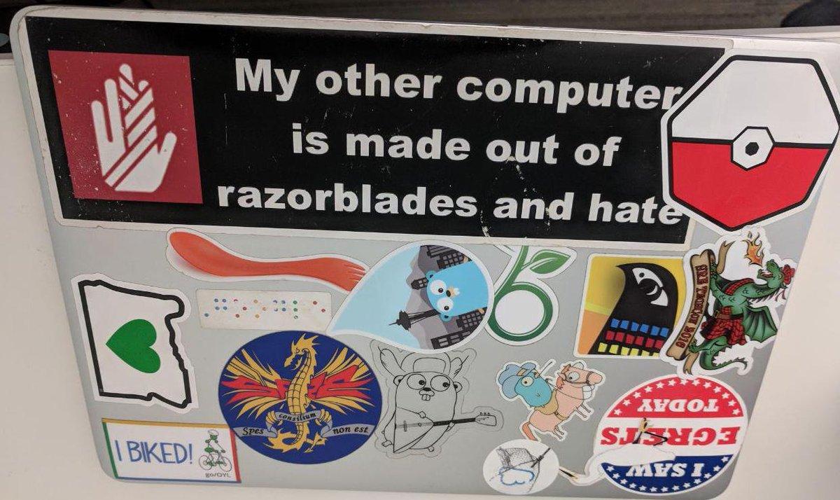 a silver laptop back full of stickers. one of them shows a black sticker with a red-white bandaged hand symbol and white text thar reads "my other computer is made out of razorblades and hate"