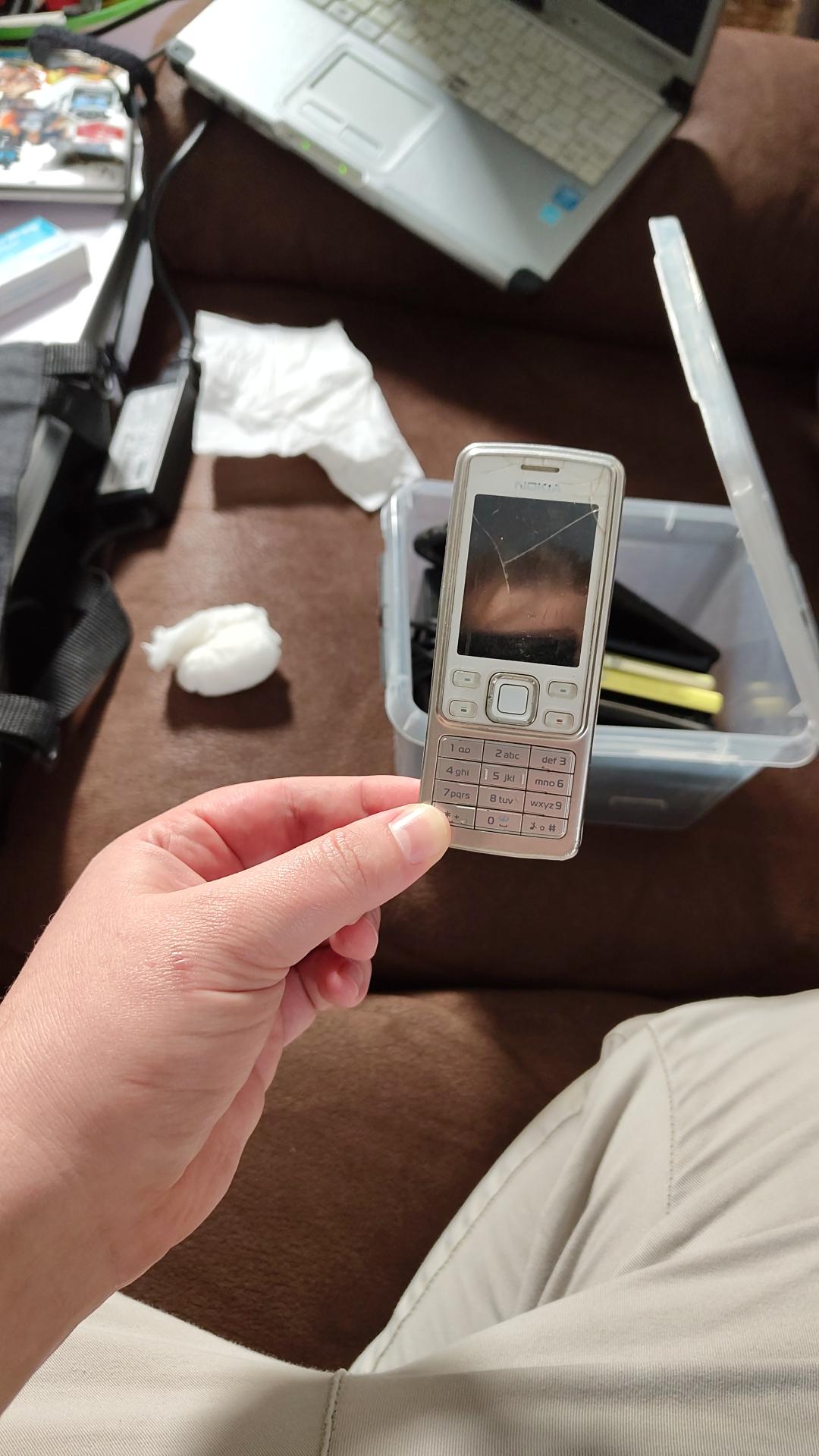 the white-silver Nokia 6300 being held between two fingers above a couch with a box with more phones.