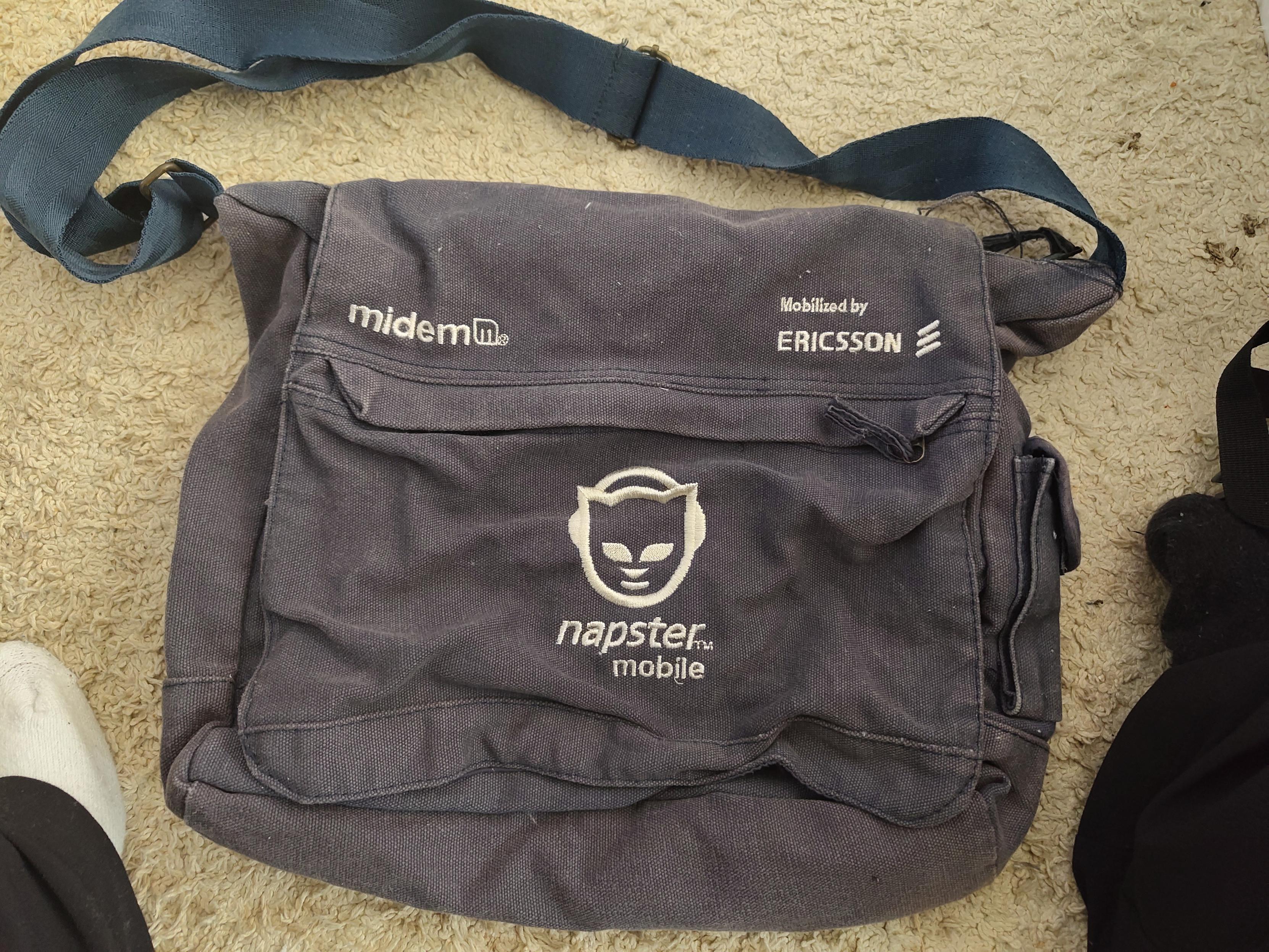 a small worn marine blue shoulder/messenger bag with the Napster Mobile logo in the middle, Midemm on the left, and Mobilized by Ericsson on the right. it's laying on beige white carpet